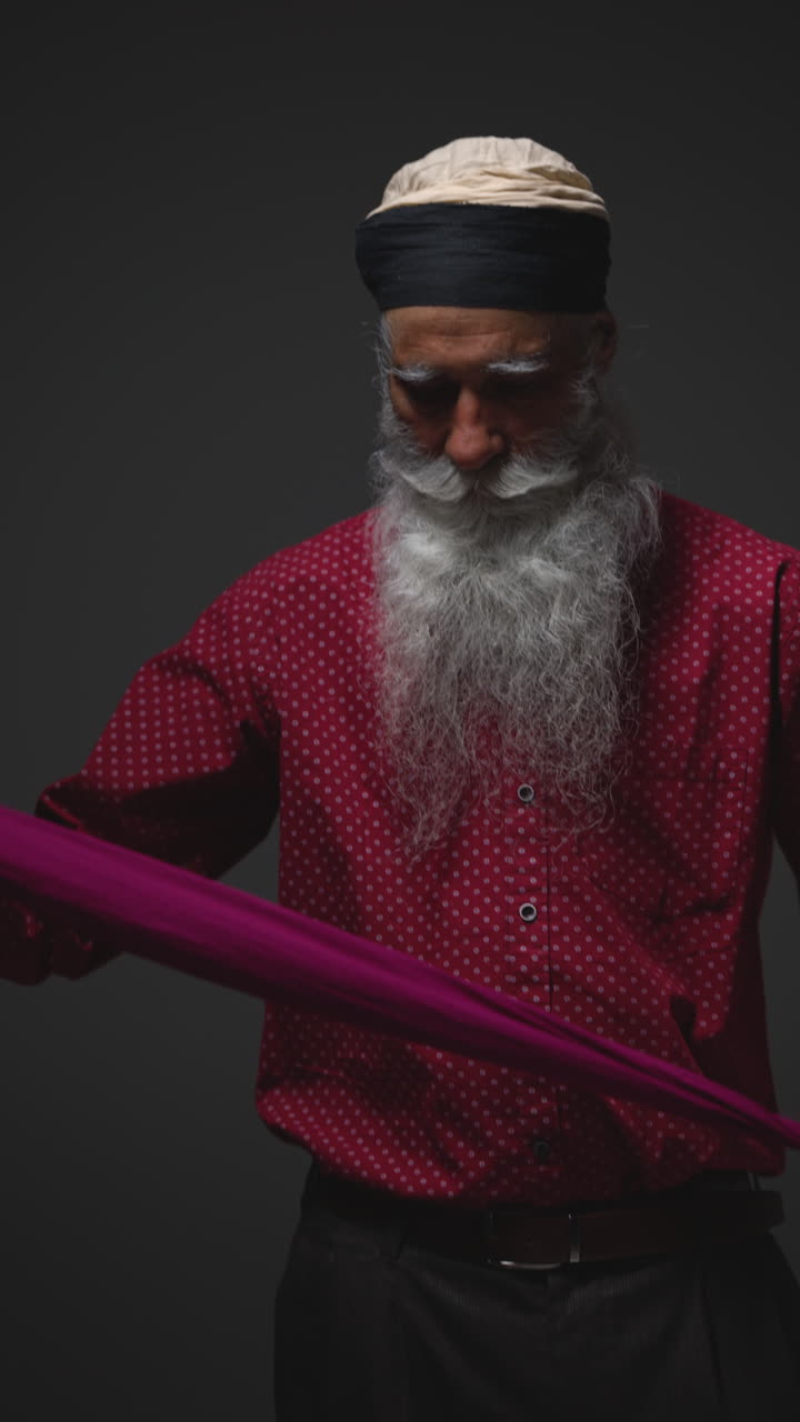 video vertical de iluminación de estudio de baja clave de un hombre sikh sénior doblando tela para turbante contra un fondo oscuro sencillo 1