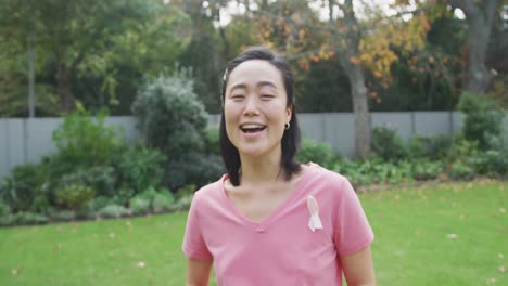 Portrait-of-happy-asian-woman-wearing-pink-t-shirt-and-cancer-ribbon,-standing-in-garden-laughing