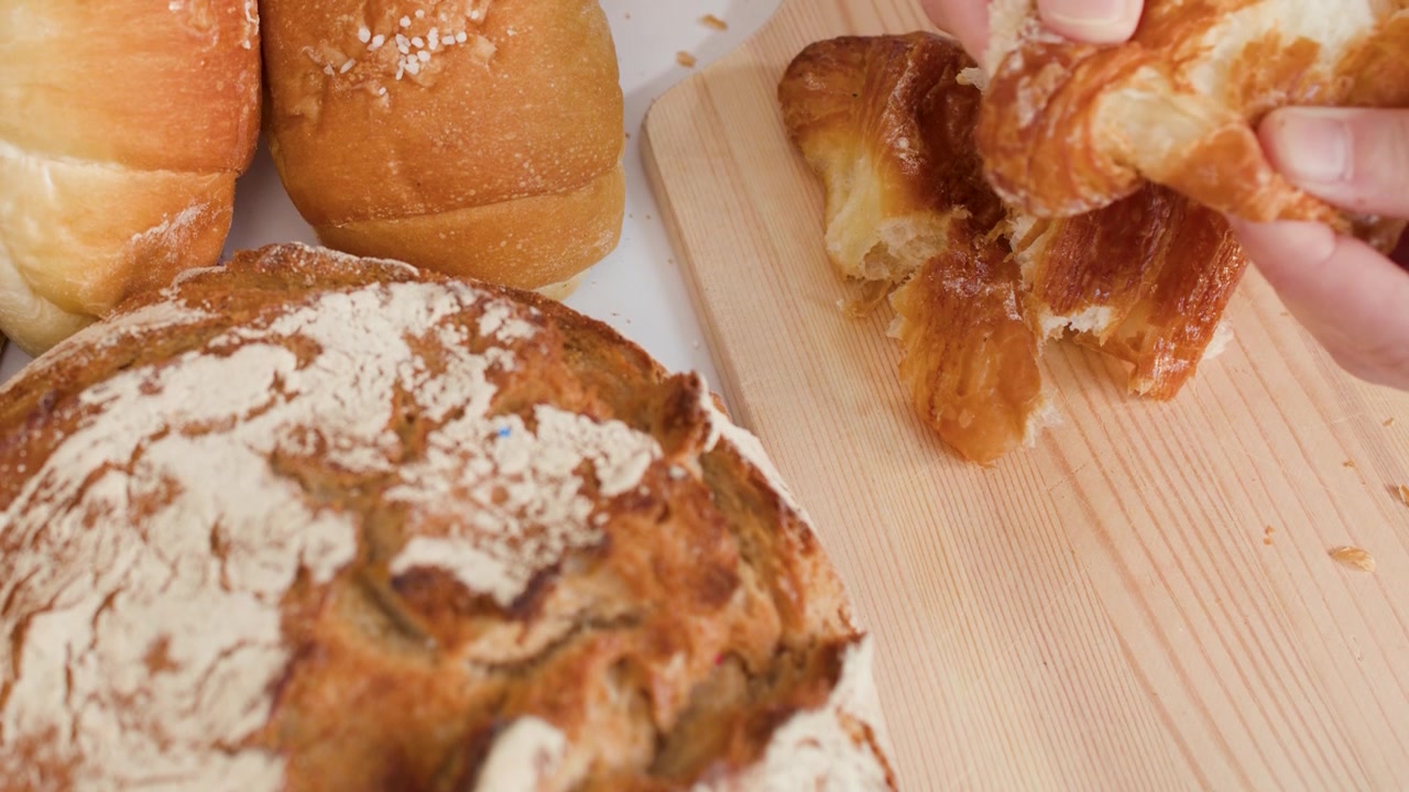 Close-up shot of hands tearing a buttery croissant on a wooden board with sourdough.