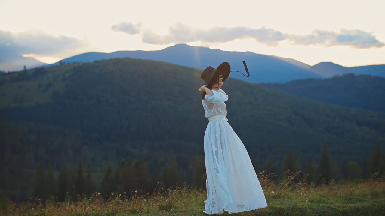 Young woman in long white dress stands on the hill holding her black hat on head. Black sting from hat fluttering in the wind. Beautiful mountainous landscape backdrop.