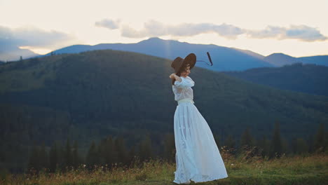 young woman in long white dress stands on the hill holding her black hat on head. black sting from hat fluttering in the wind. beautiful mountainous landscape backdrop.