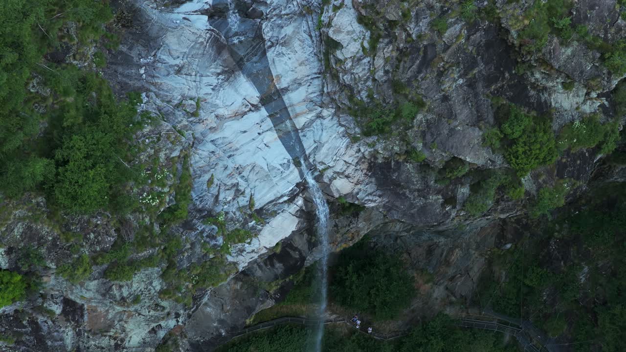 Waterfall cascading down rocky cliff face in Antrona Valley, Italian Alps, people on viewing platform at bottom. Aerial top-down view