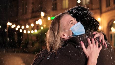 close-up view of caucasian wife and husband wearing facial masks hugging and smiling while snowing on the street in christmas