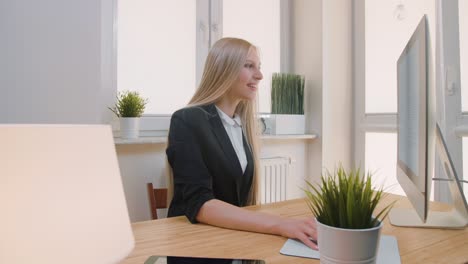 happy female working on computer in office. smiling attractive woman in business suit sitting at desk in light office and looking attentively at monitor screen.