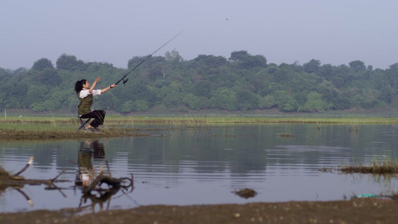 A South Asian woman casting a fishing rod while sitting on a chair by a calm lakeside, enjoying rural nature, water reflections, and peaceful daylight
