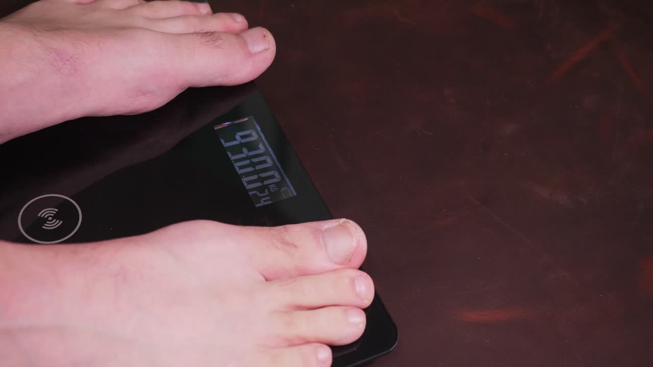 A person's hand stands on a black digital scale displaying "94.06" on a dark brown wooden floor indoors