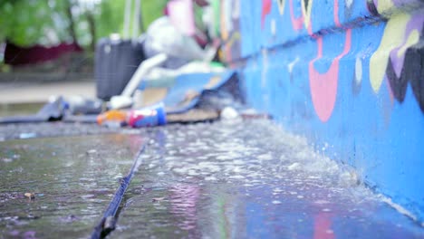 rain pouring heavily on litter next to a graffiti wall, in an urban park, creating a puddle and waterfall