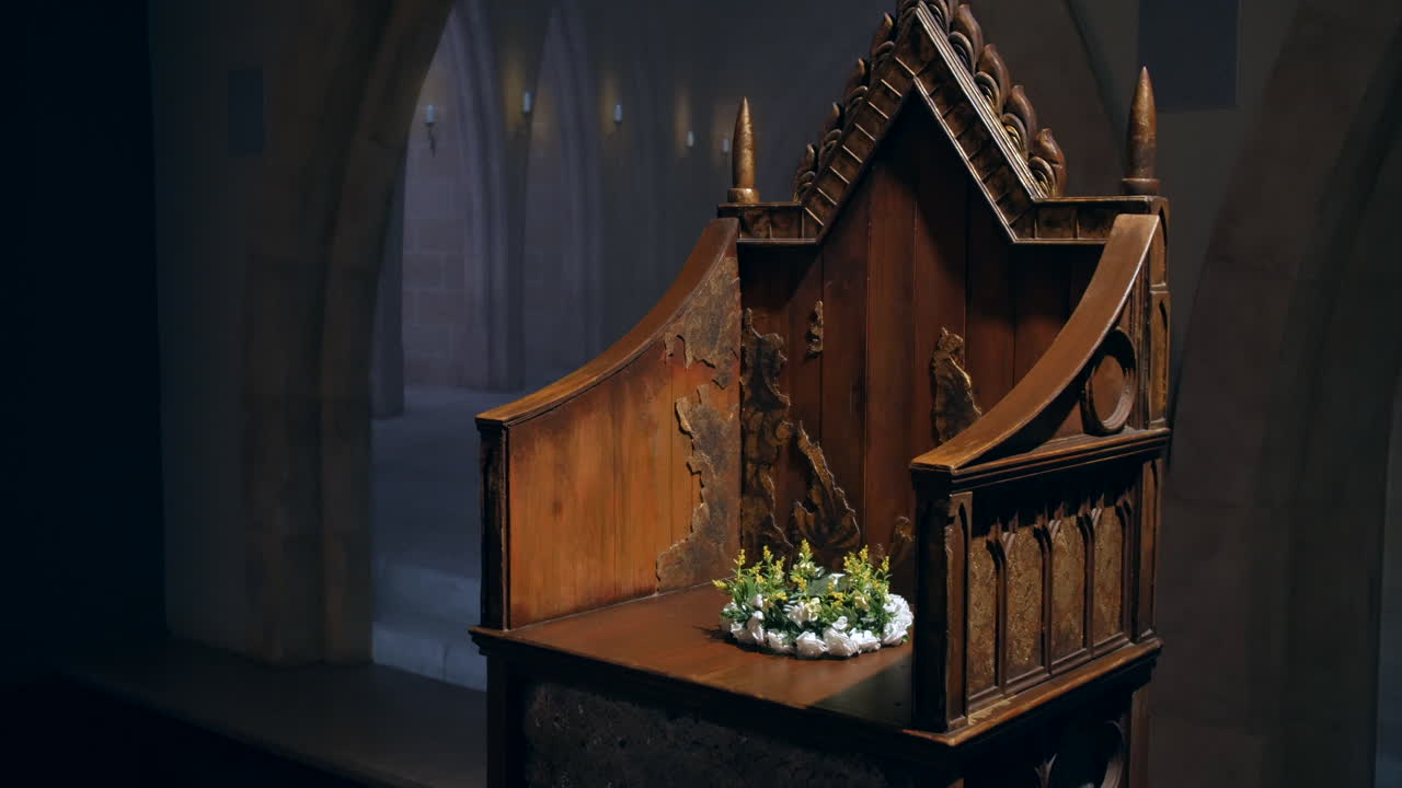 Um trono de madeira ornamentado e envelhecido com entalhes intrincados está sozinho em uma sala abobadada e mal iluminada