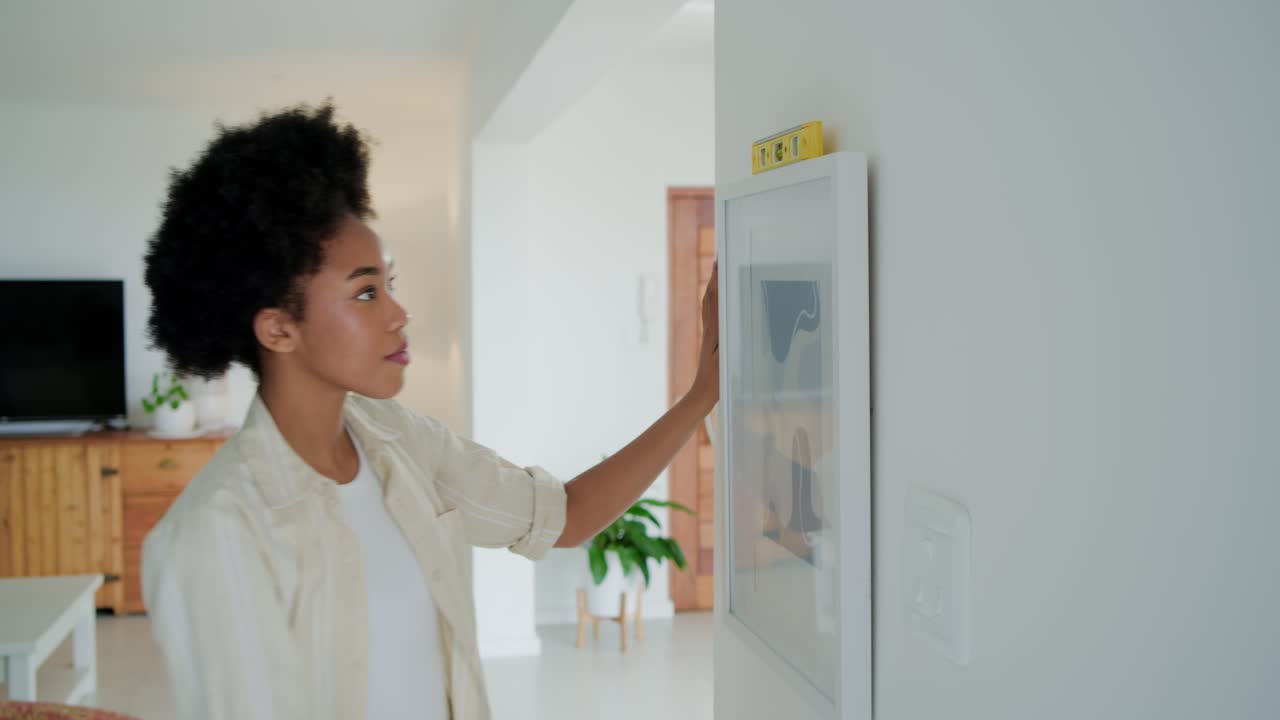 African American woman in white tee redoing frame on wall at home using level, copy space