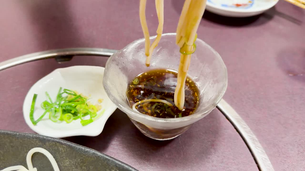 Pinze metalliche immergono noodles udon bianchi in una ciotola di vetro con salsa scura e saporita