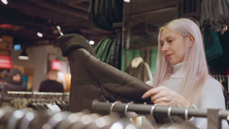 smiling fashion shopper in white turtleneck sweater lifts winter coat from rack, fingers feeling plush texture, eyes checking price tag while background boy roams store under warm lighting