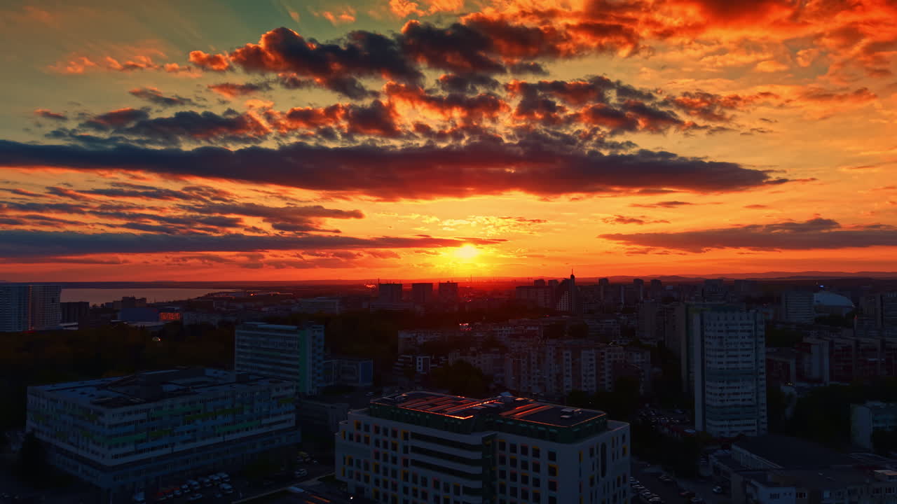 Pôr do sol sobre o horizonte de uma cidade com edifícios, nuvens e água ao fundo, durante as horas da noite.