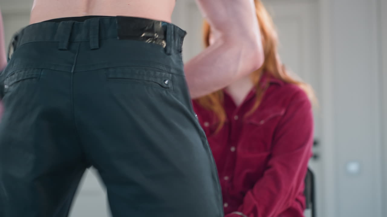 rear view bare backed male dancer in harness steps close to seated woman then slowly unfastens and lowers black trousers preparing to reveal underwear during intimate performance