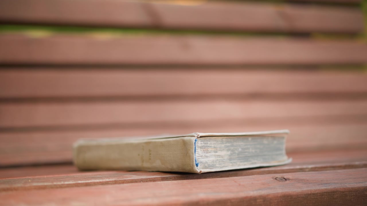 Primo piano di un libro invecchiato appoggiato su doghe di legno con sfondo sfocato. La vecchia copertina rigida mostra segni di usura, posizionata orizzontalmente su una panca alla luce naturale del giorno. Le assi di legno formano.