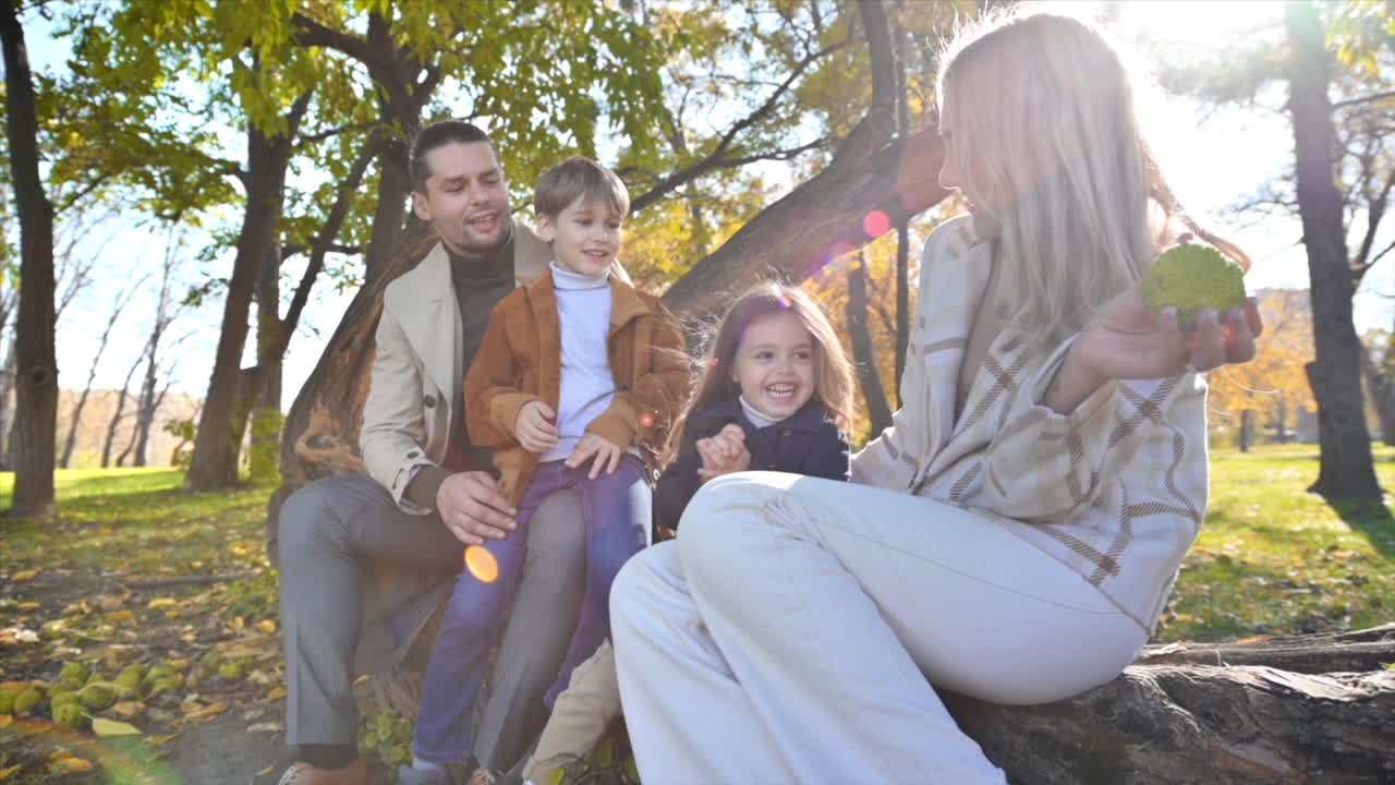 Happy family in an autumn park. Mother, father, son and daughter sitting on a tree trunk, mom playing with her daughter, yellowed trees around. Slow motion