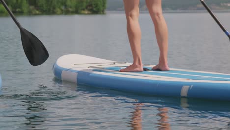 la cámara enfoca las piernas de dos chicas que están remando con la tabla y los remos en el agua