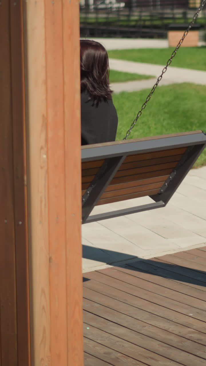 vista lateral de una mujer en una silla oscilante, su cabello soplando en el viento, en un tranquilo parque público con paisajismo detallado y personas en el fondo
