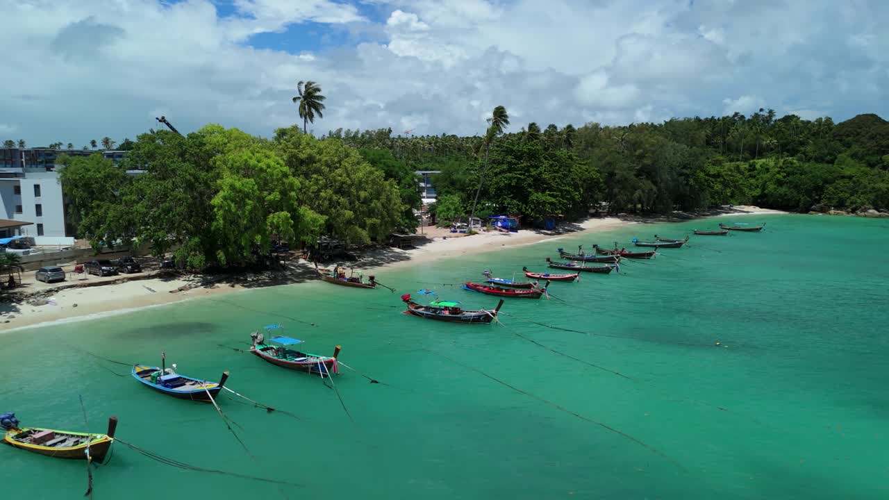 Drone pushes back over turquoise shallows as longtail boats line the shore at Rawai Beach
