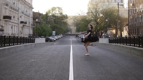 long haired young ballerina in black ballet tutu jumping cross the empty road in the european street. ballet movements. front view. slow motion