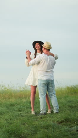 una pareja amorosa baila juntos en una colina cubierta de hierba, con el hombre con una camisa blanca, sombrero y vaqueros, y la mujer vestida con un vestido blanco y sombrero negro. la escena captura un momento tierno y alegre