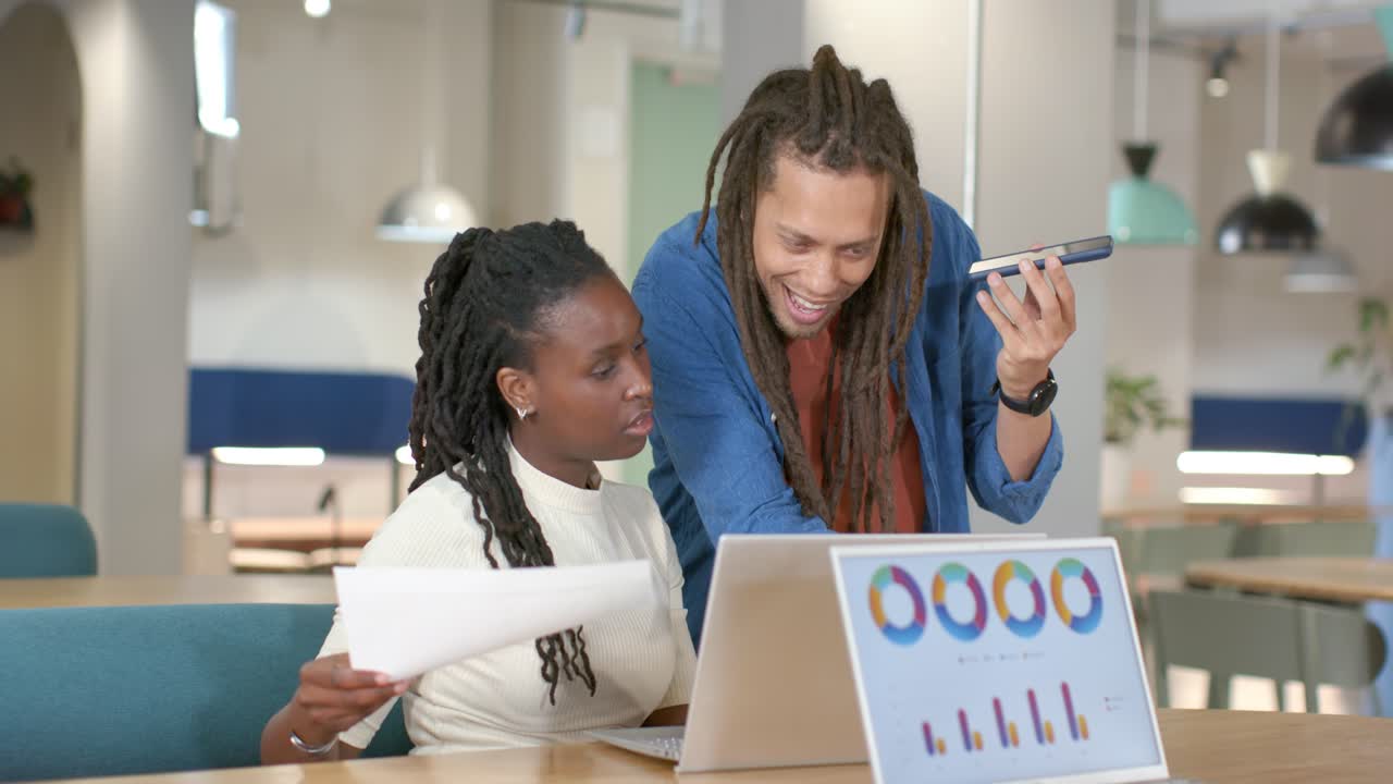African American team checking laptop charts in office, man coming with phone, woman sorting papers