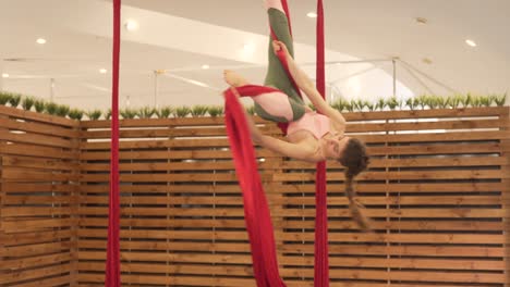 young brunette woman in a minimal and modern gym with wooden dividers doing flying yoga with some bright red hammocks, doing stunts and spinning around