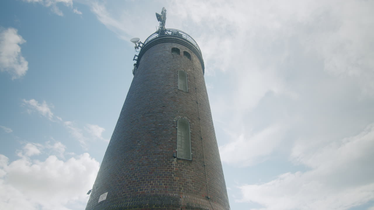 Medium wide shot of the lighthouse of Sankt Peter Ording, camera tilts down and pans to the right to reveal more of the scenery, people walking on the dam, blue sky, North Sea in the background