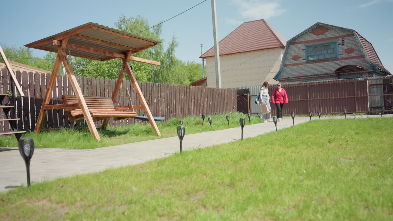 Excited ladies walk into park along paved path as one in red jacket joyfully rushes toward wooden swing under roof while others laugh and continue walking past her on sunny day