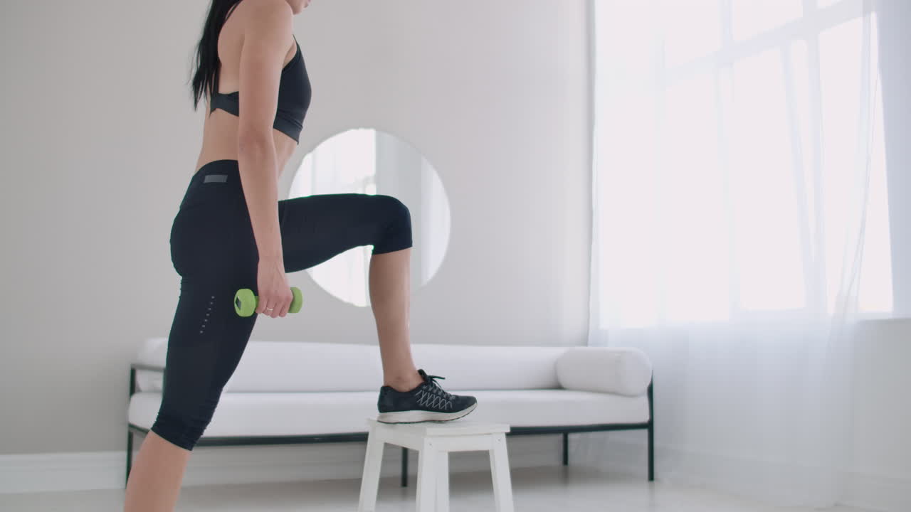 A young brunette woman performs concentrated pacing on a chair in a bright interior apartment with dumbbells in her hands.
