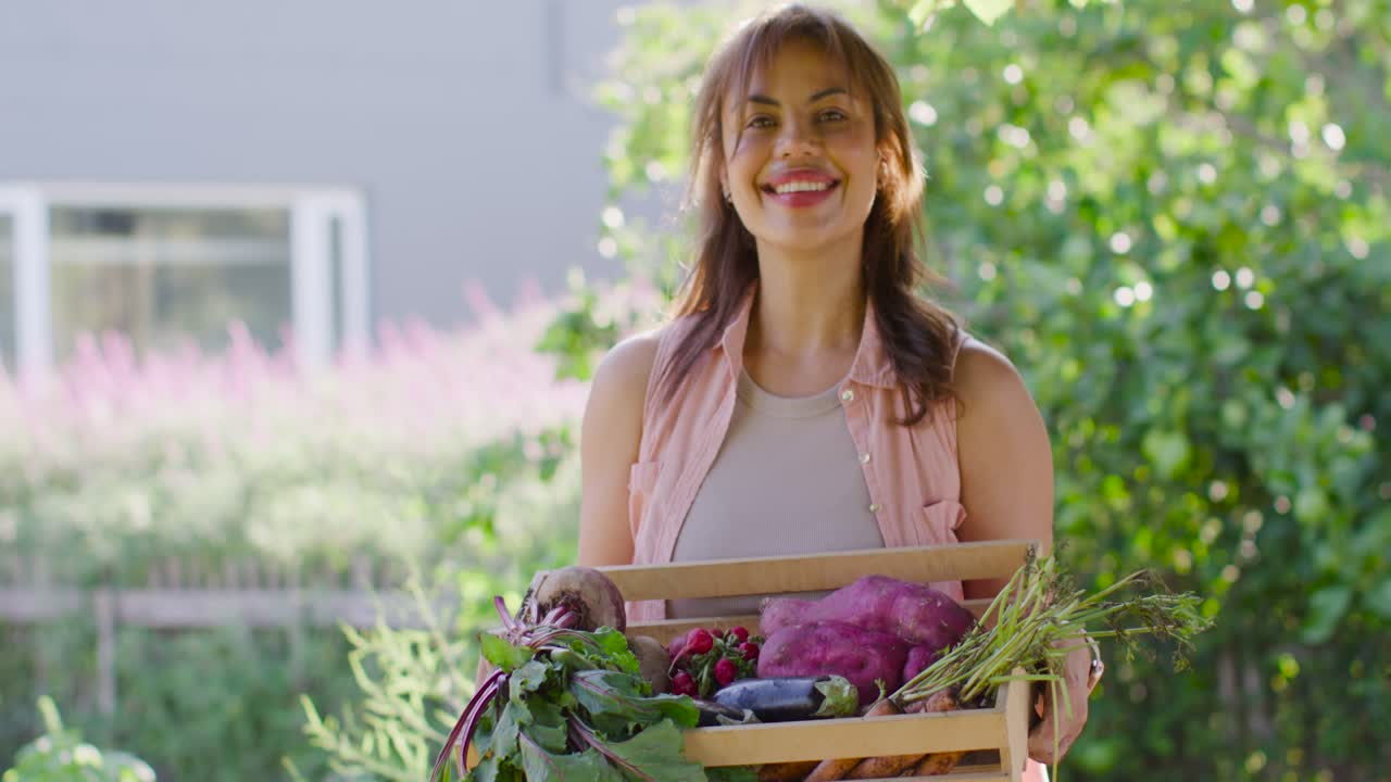 Mujer afroamericana llevando una caja de productos frescos, caminando hacia el jardín, presentando y sonriendo