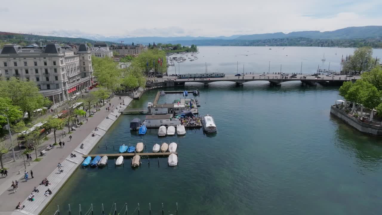 imagen de un avión no tripulado que se mueve hacia adelante que muestra el puente de la ciudad de zúrich y el lago zúrich con el metro local, banderas suizas y personas caminando a través del puente con el lago y los alpes visibles en el fondo