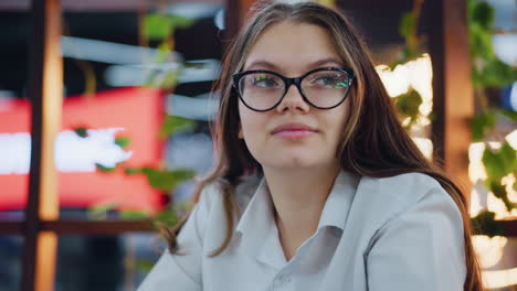 primer plano de una mujer elegante con gafas, teléfono operativo, girando a la izquierda con una mirada confiada, sentada en un bar elegante con iluminación cálida y vegetación exuberante, dedicada a la comunicación digital