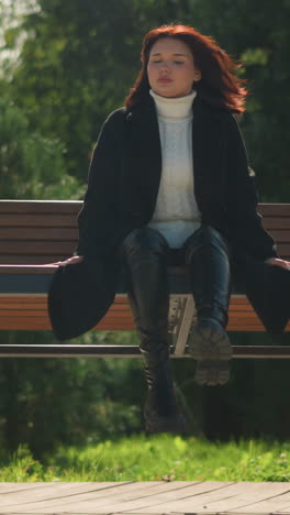 스타일리시한 검은 코트와 부츠를 입은 여자가 이 많은 공원에서 나무로 된 스에서 휴식을 취하고, 멀리서 조용히 쳐다보고, 그녀를 둘러싸고 있는 초록색으로