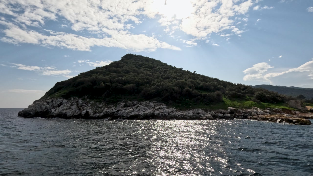 A group travels to a quiet island surrounded by water. The sun shines brightly as they get closer to the shore. Waves gently lap against the rocky coast.