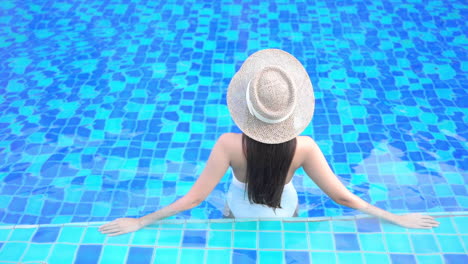 Vista-Trasera-De-Una-Mujer-Joven-Con-Sombrero-Relajándose-En-La-Piscina-Apoyada-En-Un-Borde