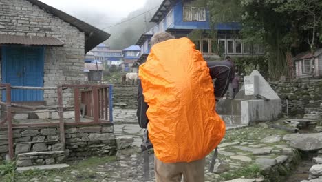 joven en cámara lenta caminando por el pueblo de nepal con mochila naranja cubierta de lluvia, annapurna