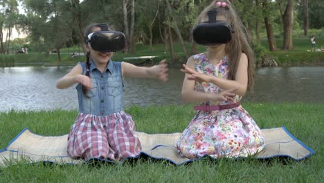 two funny little girls play in virtual reality glasses while sitting on the grass in the park on a summer evening. they wave their hands, swim in an imaginary river, and laugh. portrait. 4k.