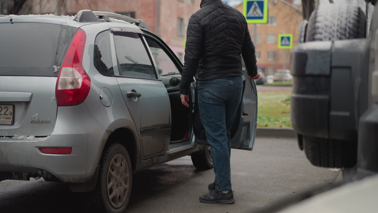 Man wearing black winter coat and blue jeans opens vehicle door and enters ash coloured car parked on urban pavement with snow droplets and water streaks visible during cold day scene