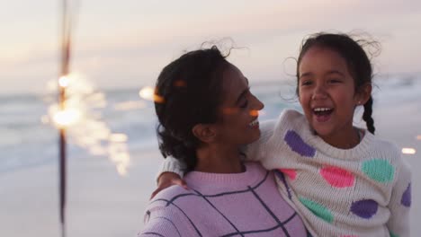Portrait-of-happy-hispanic-mother-and-daughter-playing-with-sparklers-on-beach-at-sunset