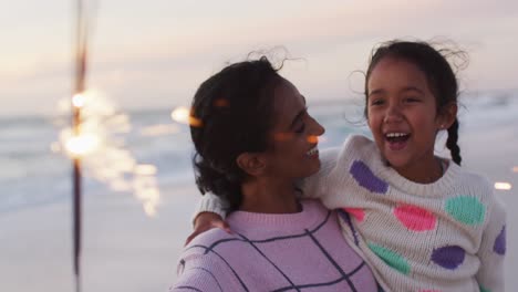 portrait of happy hispanic mother and daughter playing with sparklers on beach at sunset