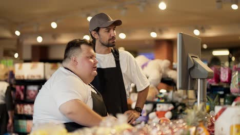 A-trainee-and-an-experienced-supermarket-worker-learns-how-to-properly-weigh-food-on-a-supermarket-scale.-Men-in-white-T-shirts-and-black-front-ones-weigh-food-in-a-supermarket