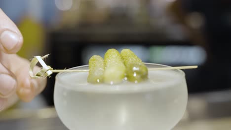 a pickle or cornicon being placed slowly on top of a martini by a bar tender