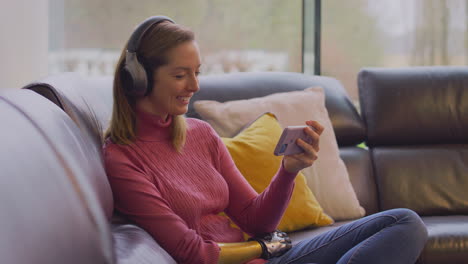 Mujer-Con-Brazo-Protésico-Usando-Auriculares-Inalámbricos-Escuchando-Música-En-El-Teléfono-Móvil-En-El-Sofá