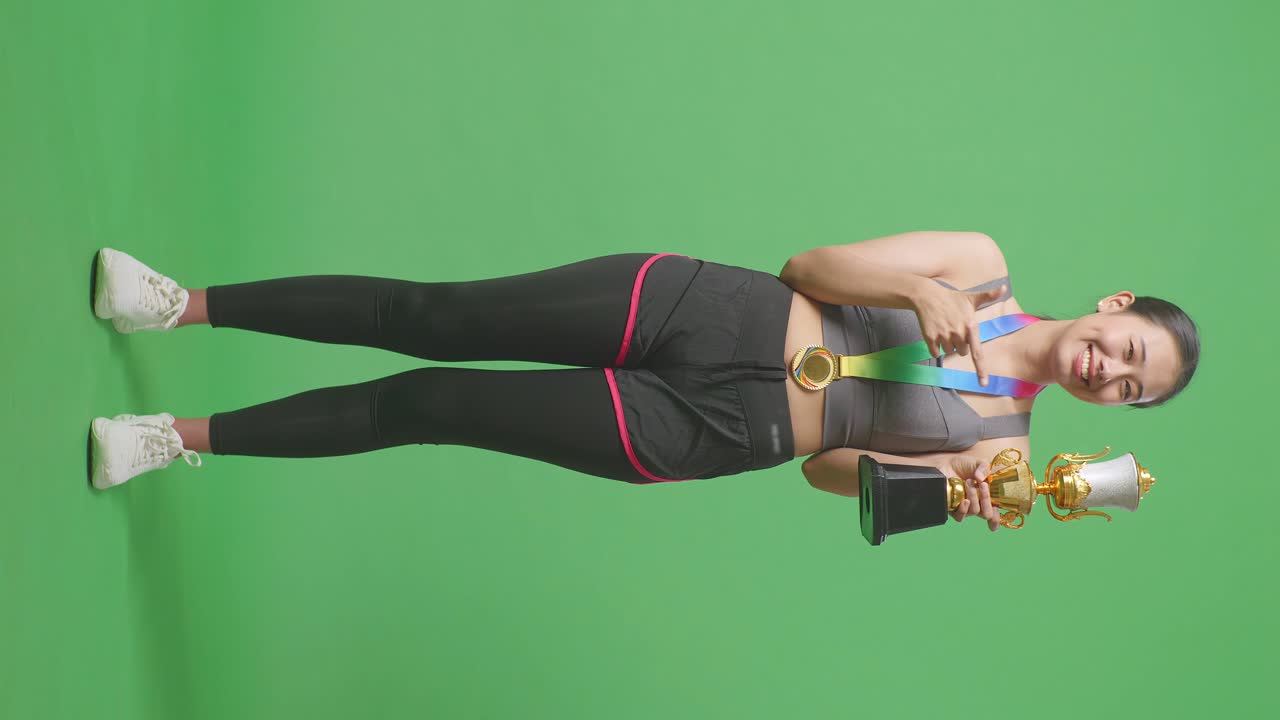 Full Body Of Asian Woman With A Gold Medal Pointing To A Gold Trophy In Her Hand Being Proud Winning As The First Winner On Green Screen Background In The Studio