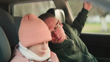 a close-up shot of a young girl dressed warmly in a pink beanie and jacket, sitting quietly in a car, with a man, likely her father, leaning in and attentively looking at something she is holding