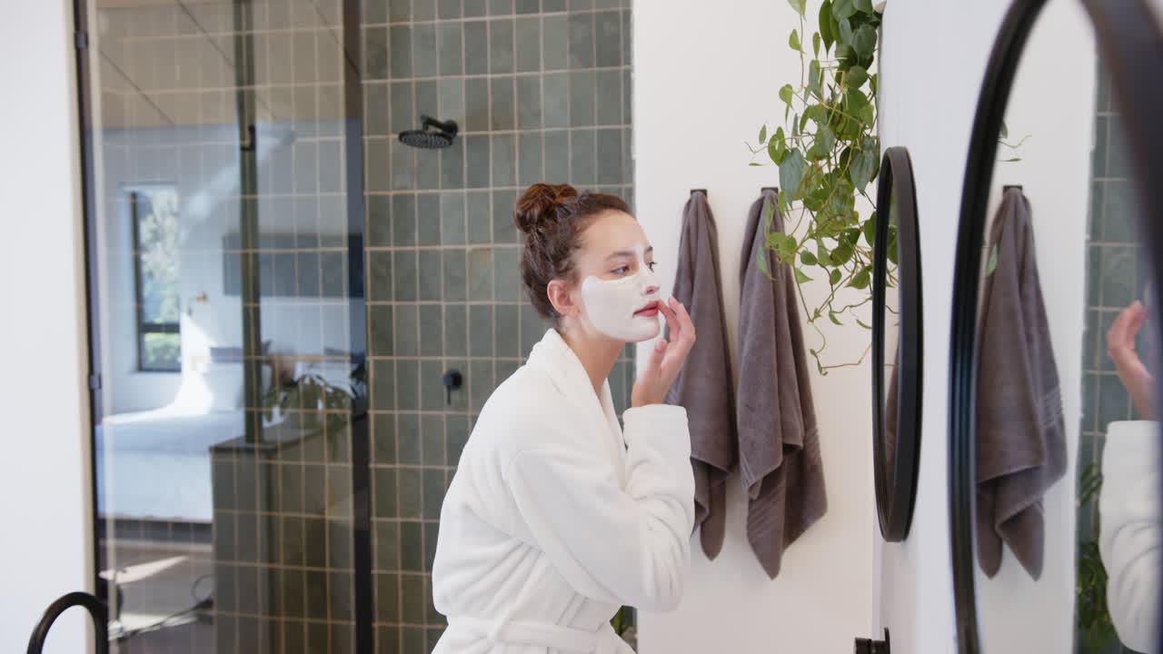 Applying facial mask, woman in bathrobe enjoying home treatment at home