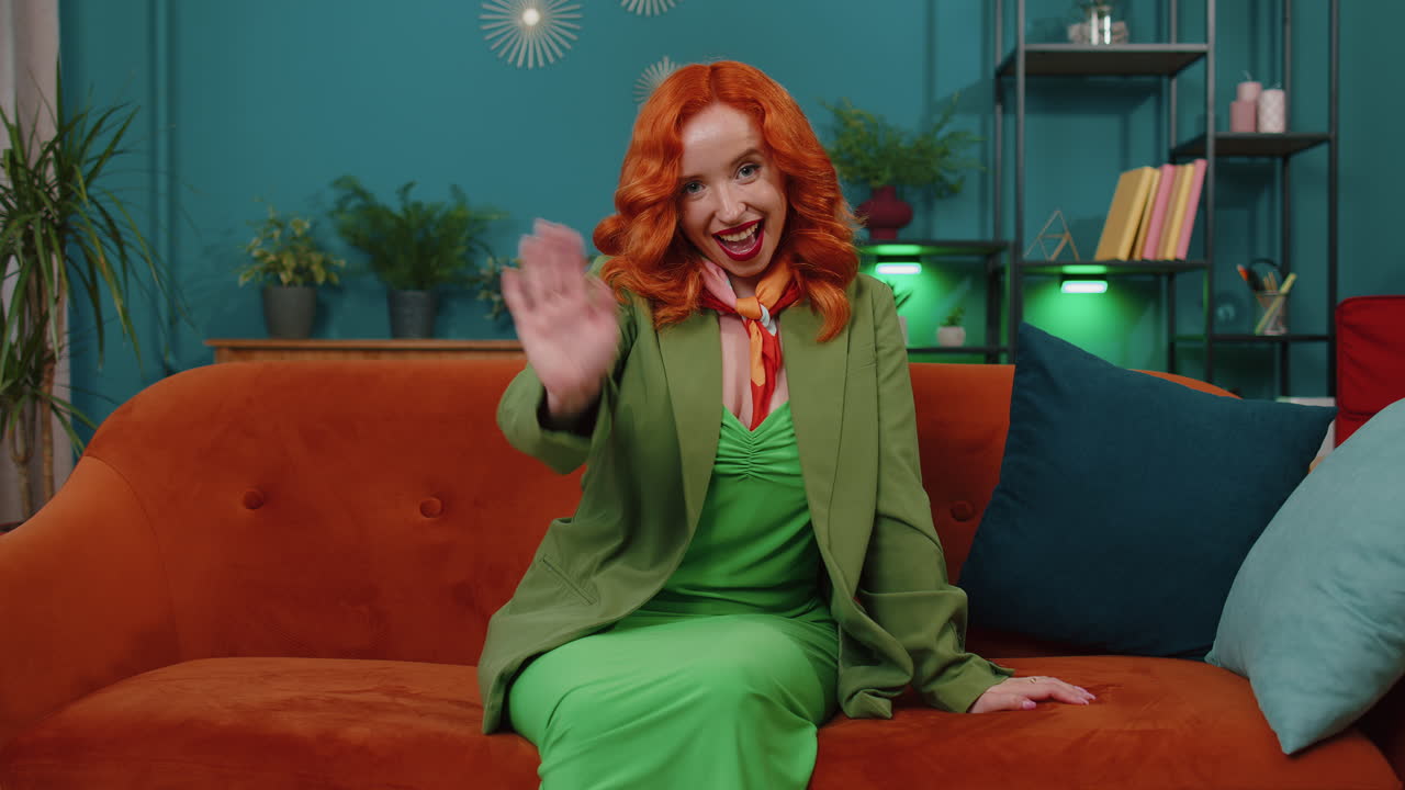 chica pelirroja sonriendo amigablemente a la cámara, agitando las manos gestando hola, hola, saludo en casa en el sofá