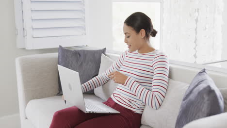 Feliz-Mujer-Birracial-Sentada-En-Un-Sofá-Usando-Una-Computadora-Portátil-Para-Comprar-En-Línea,-Cámara-Lenta