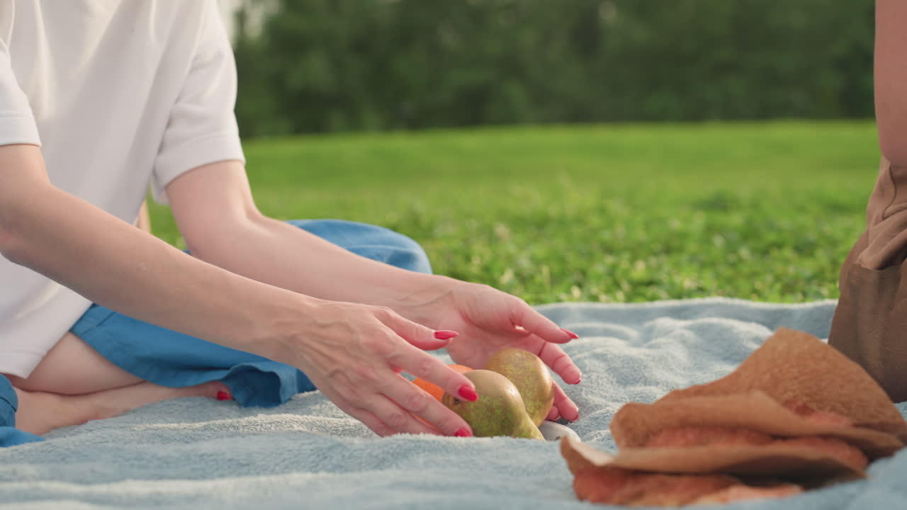 lower view of park goers seated on picnic blanket, one handing fruit to other while sandwich lies nearby, children running around on green grass, summer bonding moment of sharing