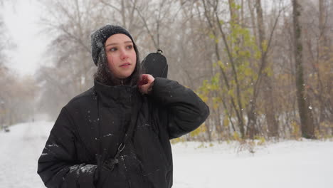 white chic in winter jacket adjusts her guitar in its pack, gazing thoughtfully into the distance, with a snow-covered bench in the background amidst falling snow